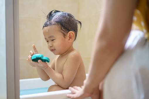 Adorable Asian little boy playing with bath toy in bathtub while his mother bathing him.