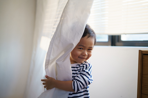 Adorable curious Asian baby boy playing hide and seek with his parent hiding behind white curtain in bedroom happily.