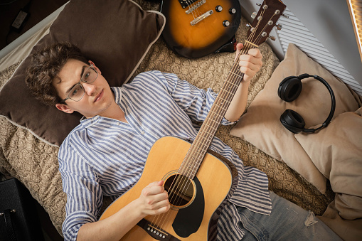Portrait teenager man playing acoustic guitar lying on comfy bed relaxing enjoy art entertainment hobby top view. Male guitarist musician player singing song music instrument resting at home