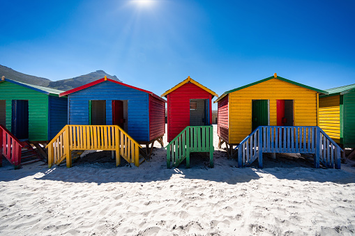 Muizenberg Beach close to Cape Town. Colourful beach with beach huts side by side in a row on the white sandy beach of Muizenberg under blue cloudless summer sky backlit from the summer sun. Muizenberg Beach, Muizenberg, Cape Town, South Africa, Africa