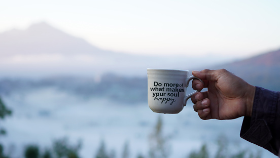 Inspirational quote - Do more of what makes your soul happy. With person holding cup of coffee on foggy mountain background. Happiness, self love and care concept. Healthy lifestyle.