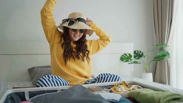 Happy young Asian woman enjoy packing clothes in suitcase for vacation trip. Cheerful energetic happy woman dances while packing suitcase for travel on bed in bedroom. Travel concept