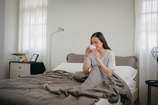 In the morning, a smiling Asian woman sits in bed, enjoying a morning coffee.