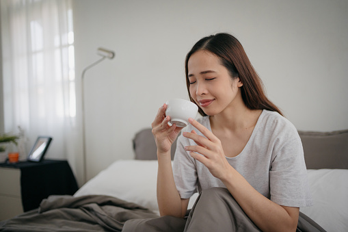 In the morning, a smiling Asian woman sits in bed, enjoying a morning coffee.
