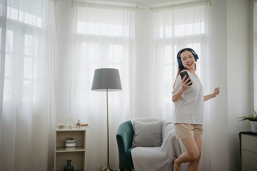A joyful Asian woman, wearing headphones, dances to her favorite music in the bedroom.