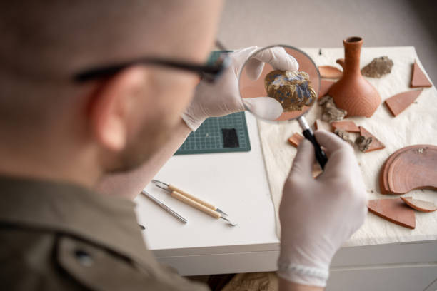 Archeologist work in office, studying newly discovered ancient civilization cultural artifact, gold coins in soil, fossil remains trough magnifier glass