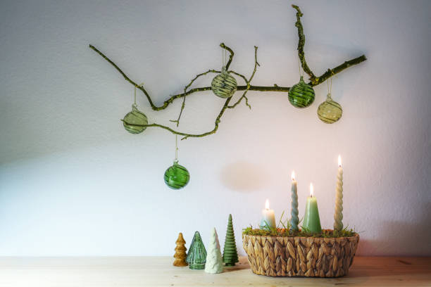 istock Advent and Christmas decoration with four different candles in a basket and small artificial trees under a hanging bare branch with green glass balls against a light wall, copy space 1753644503