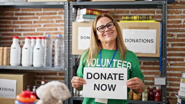 Middle age hispanic woman volunteer holding donate now message on paper at charity center