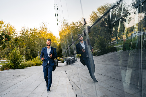 Businessman using phone in front of a corporate building and drinking coffee outdoors. Going to work in the morning.