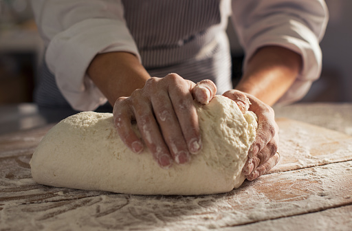 Closeup shot of baker's hands kneading dough in the early morning light. Natural and organic dough made from flour and yeast on wooden table. Woman hands knead dough to make fresh bread loaf at bakery.