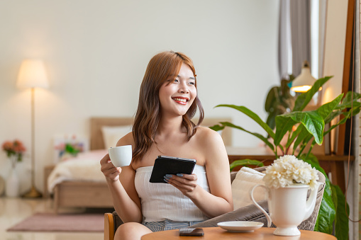 From the comfort of her home, a young Gen Z Asian transgender woman enjoys authentic interactions with friends on social media using digital apps. She takes a break, sipping tea while staying digitally connected with her digital tablet in her cozy and inviting environment.