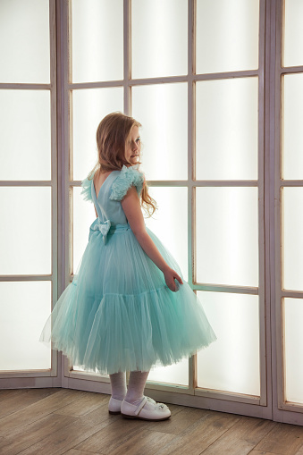 Rear view of sad cover girl in art azure dress posing at large window, full length, showing excitement. Studio shot unhappy pensive stylish kid lady. Children emotion concept. Copy ad text space