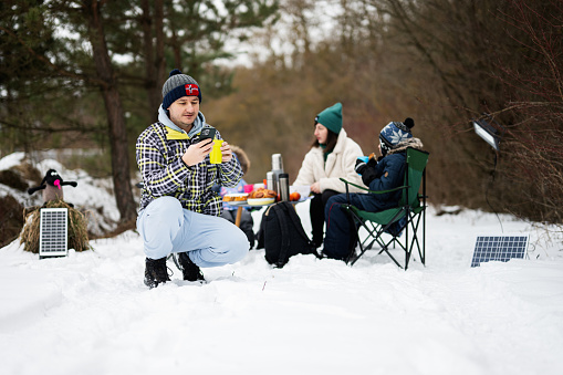 Man with phone at hand against his family in winter forest spending time together on a picnic.