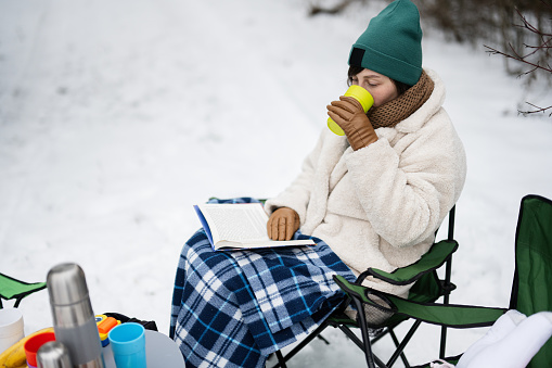 Woman sit in chair, read book and drink tea in winter forest on a picnic.
