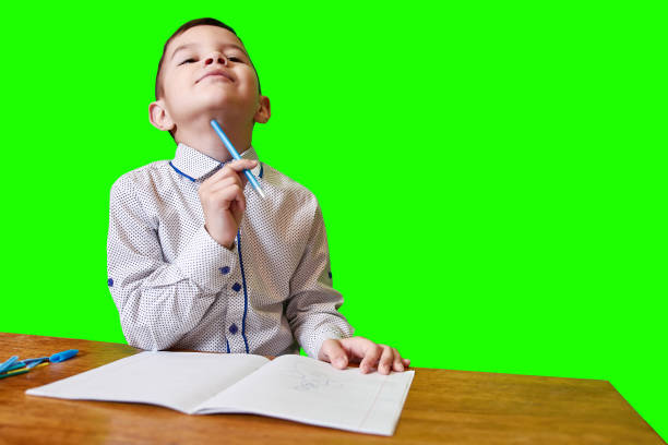Cute pensive boy holding a pen in his hand sitting at a table with an open notebook on a green background.