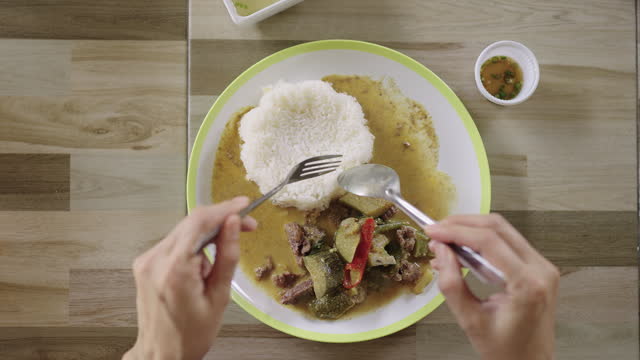 Top view of a person's head enjoyably eating Thai green curry with beef and eggplant with steamed white rice using spoon and fork at a cafeteria.