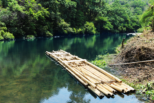 Photo of a bamboo raft floating on the river. This bamboo raft is made from bamboo woven together. It has a rectangular shape. The floor of the raft is covered with bamboo. On top of the raft there is a roof covered with leaves.