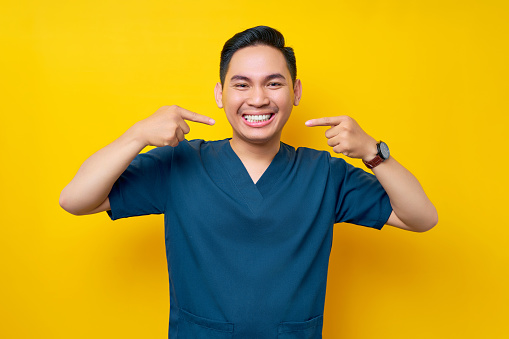 Professional young Asian male doctor or nurse wearing a blue uniform showing well-groomed white teeth isolated on yellow background. Healthcare medicine concept