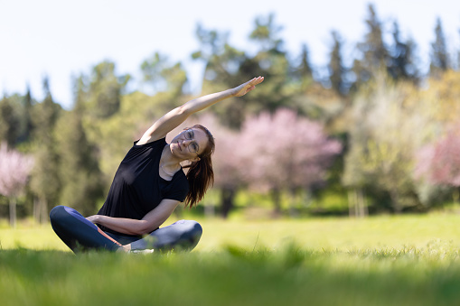 An adult woman sitting in lotus position in blooming park and bending to the side. Mid shot