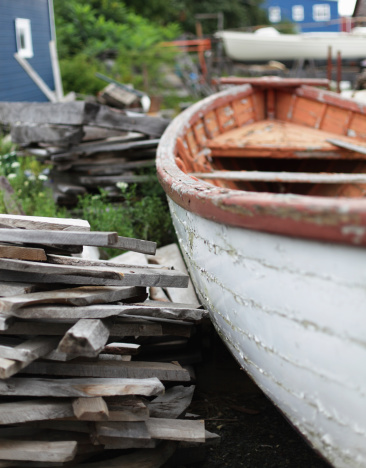 An aging, rotting dory with peeling paint is sitting behind a Lunenburg, Nova Scotia, Canada boat building shop next to piles of randomly stacked raw timber. Extremely shallow depth of field with selective focus on the midsection gunwale of the boat. The coastal town of Lunenburg is a UNESCO World Heritage Site.
