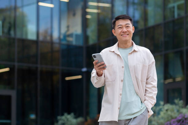 istock Young Asian man businessman, freelancer, designer standing outside office buildings, using mobile phone and smiling at camera 1753801117