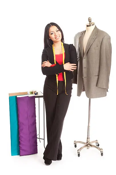 Subject: Asian woman dressmaker, fashion designer, seamstress, or tailor standing confidently with arms crossed, smiling for the camera in a small business environment. A tape measure drapes from her neck. By her side, bolts of fabric lean against a small table with spools of thread sitting on it, and next to her a suit jacket hangs on a mannequin.