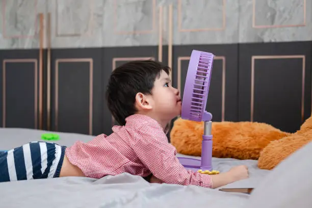 Photo of Asian boy Lying on the mattress on a hot day Playing with a portable fan happily