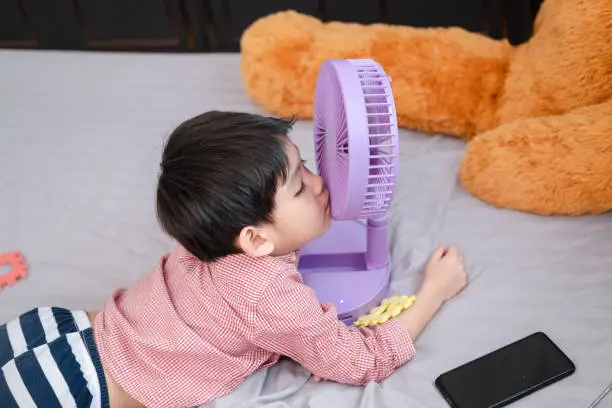 Photo of Asian boy Lying on the mattress on a hot day Playing with a portable fan happily