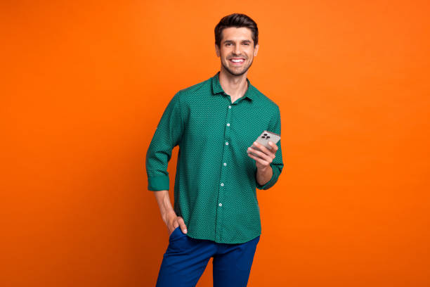 Photo of cheerful young man brunet hair hold new apple iphone gadget chatting broker working online isolated on orange color background stock photo