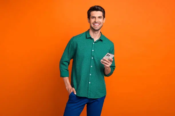 Photo of cheerful young man brunet hair hold new apple iphone gadget chatting broker working online isolated on orange color background.