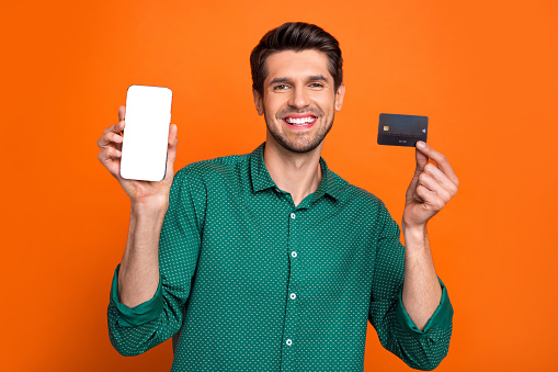 Photo of young promoter man wear green shirt hold debit card payment smartphone screen finance app isolated on orange color background.