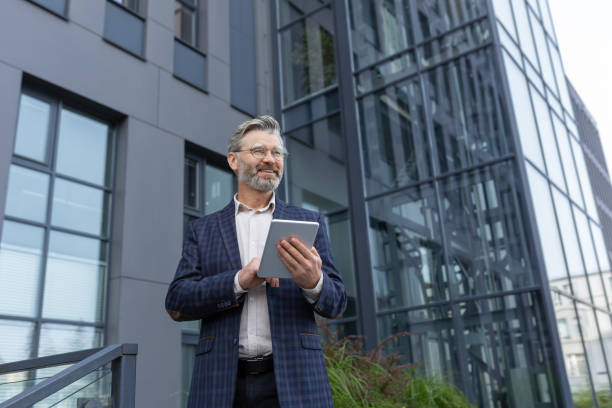 istock Successful gray-haired businessman smiling and happy, man using tablet computer, senior boss reading news and browsing online pages from outside office building walking around the city 1753870861