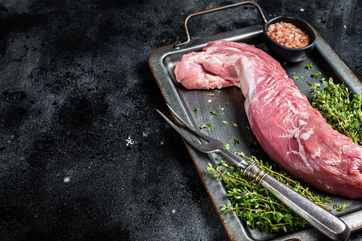 Raw pork tenderloin fillet meat in steel tray with herbs. Black background. Top view. Copy space.