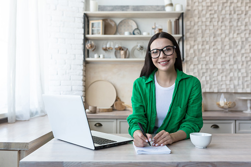 Retrato de una joven estudiante que estudia a distancia con un ordenador portátil. Sentado en la cocina a la mesa con un cuaderno y sonriendo mirando a la cámara photo