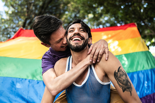 Gay couple piggyback outdoors with rainbow flag on background