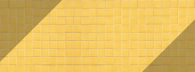 Yellow square tile wall background with sunlight and shadow on surface in panoramic view.