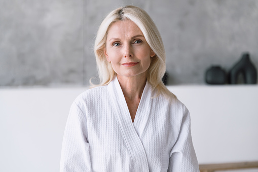 Portrait of middle aged woman in white shower robe with healthy skin face doing beauty procedures and anti-aging treatment at home bathroom. Self care and morning routine concept.
