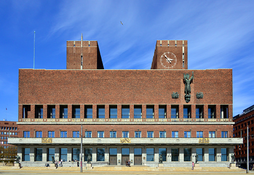 Oslo, Norway: Oslo City Hall (Oslo rådhus) - built 1931–1950 designed in functionalist style by the architects Arnstein Arneberg and Magnus Poulsson. The town hall is located in the center of Oslo on the north side of Pipervika, the south façade faces Rådhusplassen, the town hall piers (Rådhusbryggene) and the Honnørbryggen pier by the Oslo Fjord. It is the venue for the yearly awarding of the Nobel Peace Prize. The city's coat of arms central figure is St. Hallvard, shown on the façade.