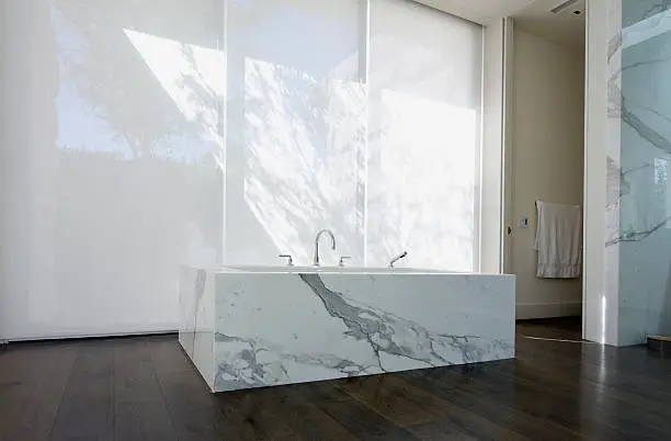 "Low angle view of a modern bathroom with stand-alone marble clad tub, hardwood floor and vertical shades."
