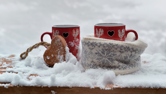 two red mugs with heart shaped one in a woolen hat  on the snow- valentine concept