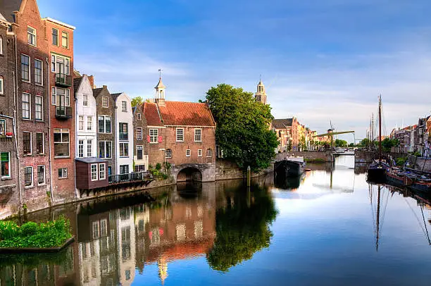 "Historic cityscape along a channel in Delfshaven, a district of Rotterdam, the Netherlands. Visible are typical dutch architecture, historic sailing boats,windmill, restaurants, colorful reflection in the river, blue and dramatic cloudscape and beautiful sunset atmosphere."