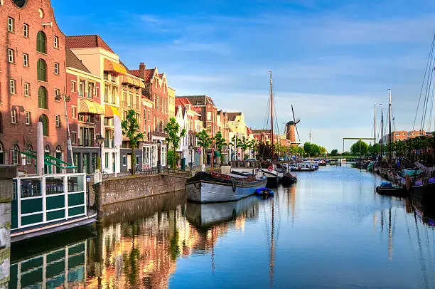 "Historic cityscape along a channel in Delfshaven, a district of Rotterdam, the Netherlands. Visible are typical dutch architecture, historic sailing boats,windmill, restaurants, colorful reflection in the river, blue and dramatic cloudscape and beautiful sunset atmosphere."