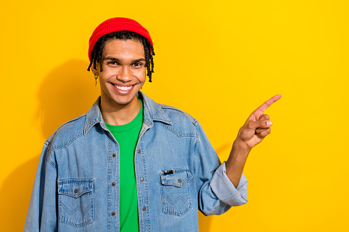 Photo of cool positive guy dressed denim jacket beanie pointing empty space isolated yellow color background.