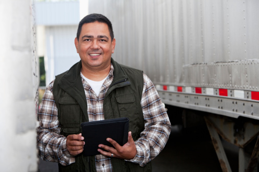 Hispanic truck driver (40s) standing next to semi-truck, holding digital tablet.
