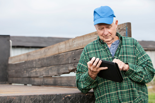 A senior man, in his 60s with white hair under his blue hat, is working on a farm, using a dgital tablet.  He is leaning against the back of an old work truck used for hauling crops and farming equipment.  He is looking down and using the touchsreen.