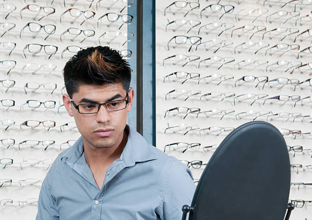 Attractive young hispanic man trying on glasses in front of a mirror at the optometrist's office.  Rows of eyeglass frames blurred in the background.Click below for more of my OPTOMETRY images: