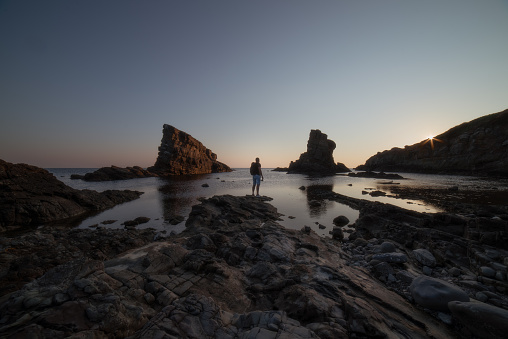 Young adult male is watching the sunset or sunrise by the rocky sea shore. Big rock formations in the sea in front of him