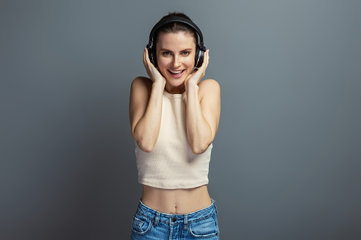 Young cheerful woman enjoying music with headphone, wearing casual top, isolated on grey studio background