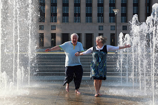 a mature / senior couple hold hands and dance in celebration in some sunlit fountains