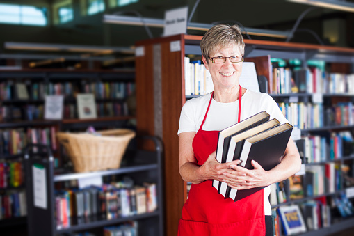 Cute volunteer librarian standing confidently holding books in library with rows of books in background